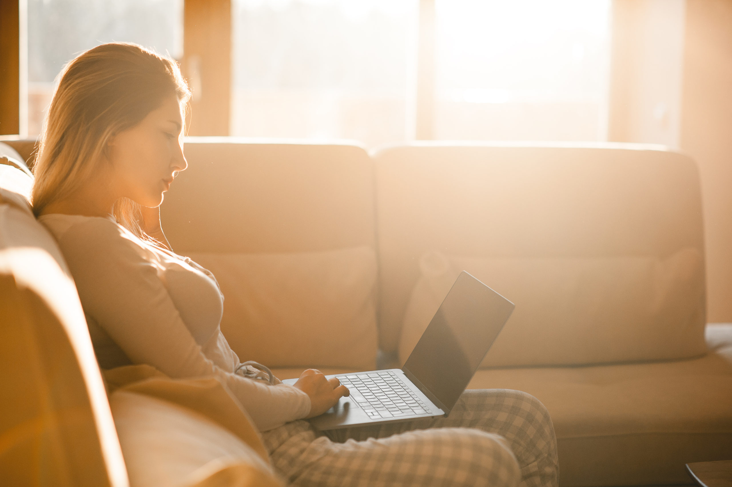 Woman sitting on her couch on her laptopped, bathed in sunlight.