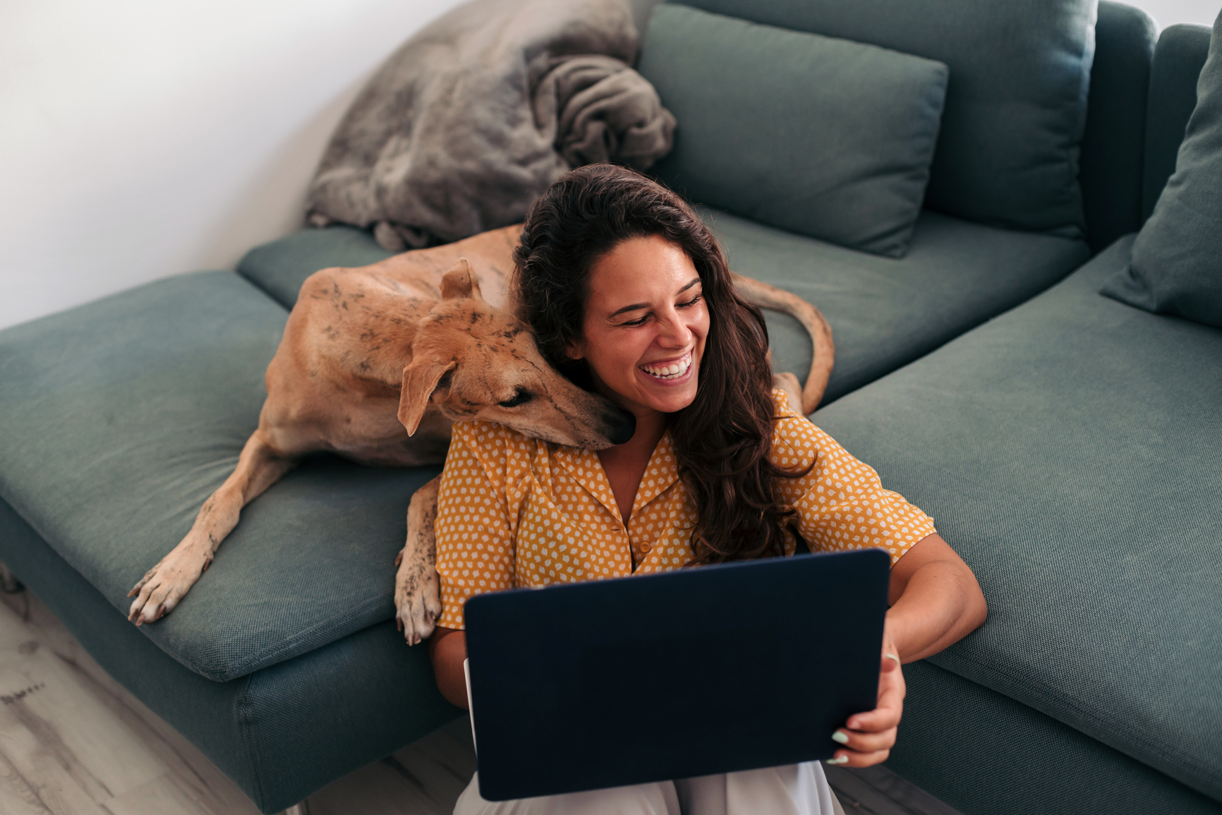 Woman sitting against her couch, holding a laptop, with her dog's head on her shoulder.