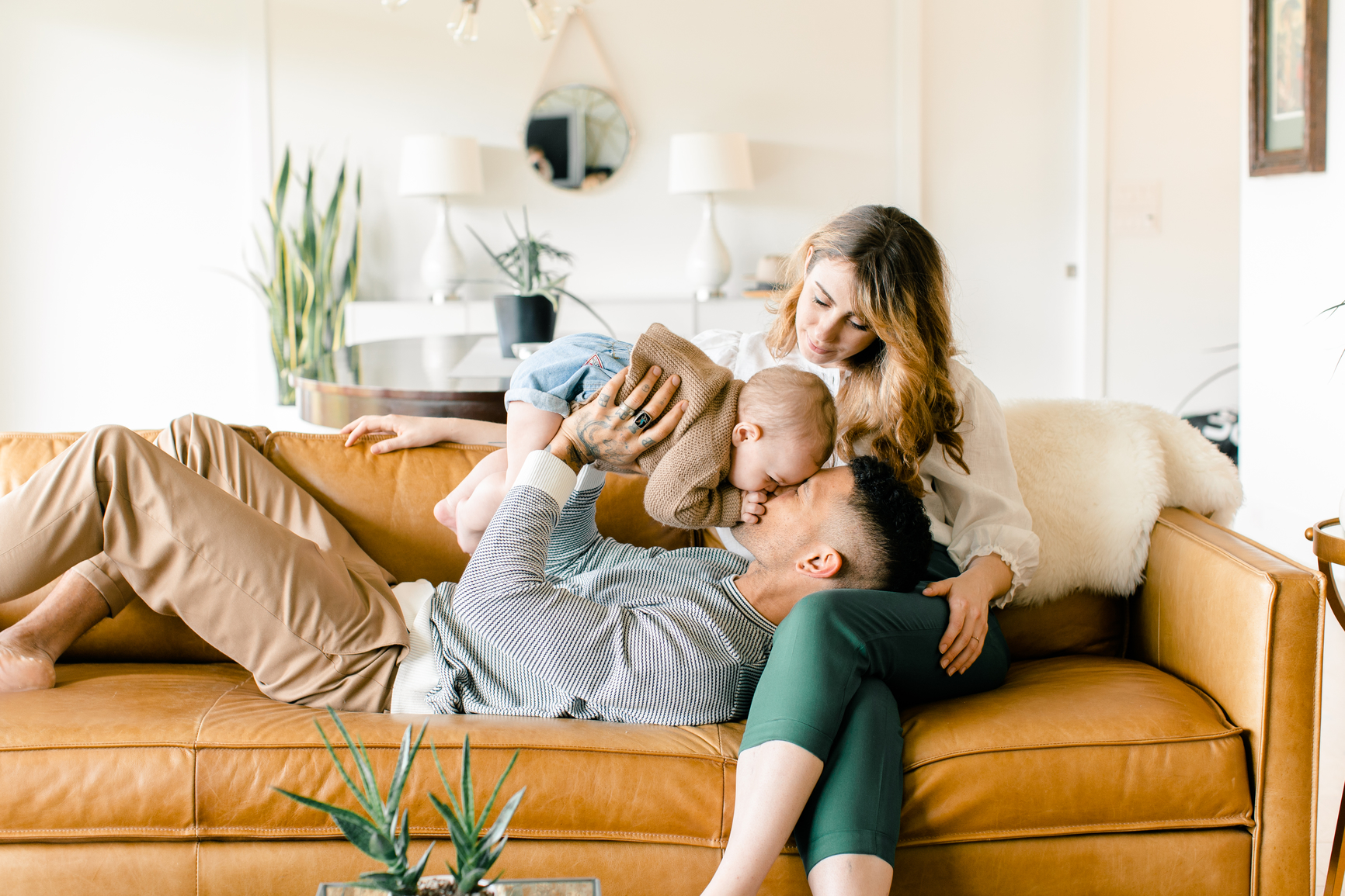Picture of a young family on their couch. The husband is playing with a baby, and the wife is working on her laptop.